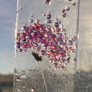 K&ouml;nnte beinhalten: Ein Glasornament mit einer lila und pinkfarbenen Blume auf transparentem Hintergrund. Die Blume besteht aus kleinen Glasperlen und hat einen zarten Stiel. Das Ornament h&auml;ngt an einem lila Band.