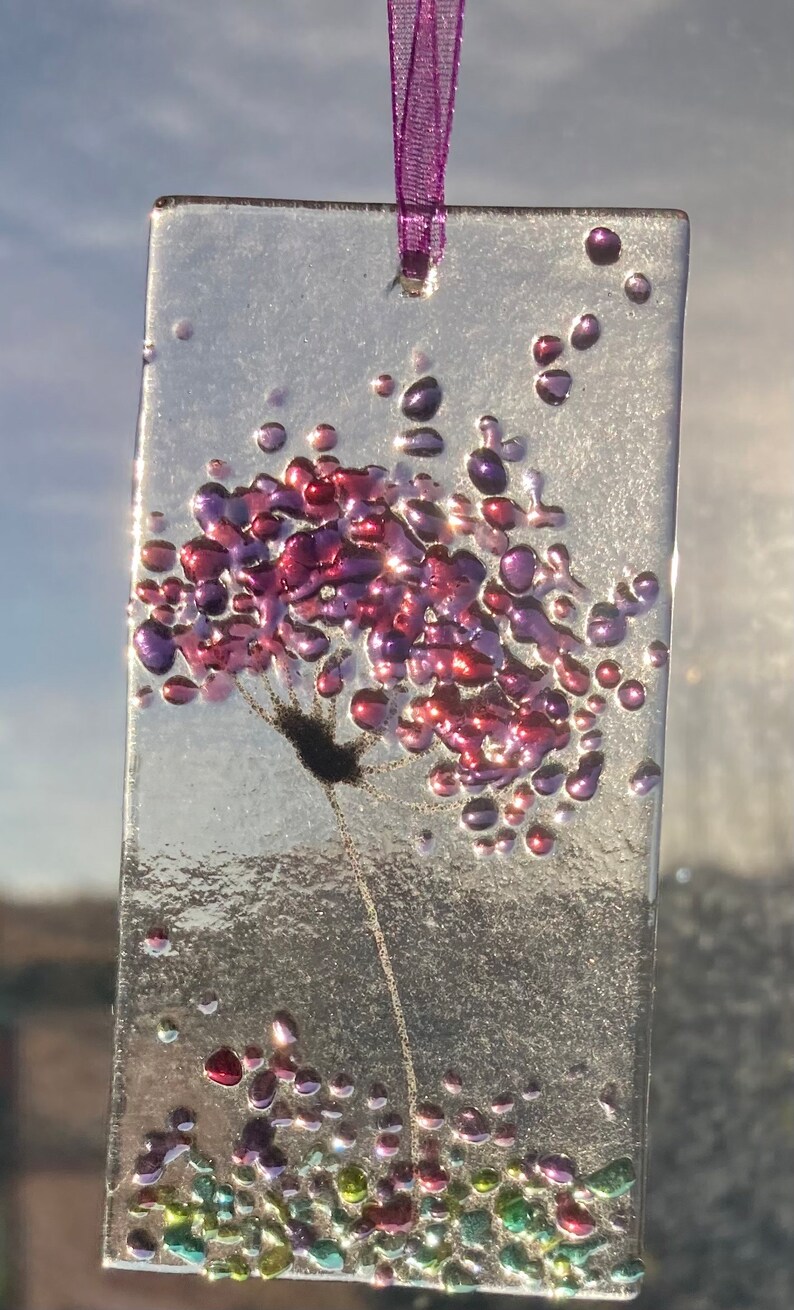 K&ouml;nnte beinhalten: Ein Glasornament mit einer lila und pinkfarbenen Blume auf transparentem Hintergrund. Die Blume besteht aus kleinen Glasperlen und hat einen zarten Stiel. Das Ornament h&auml;ngt an einem lila Band.