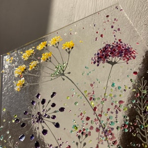 May include: A glass art piece featuring a floral design with yellow, purple, green, and red glass pieces. The flowers are arranged in a cluster and have a delicate, whimsical look.