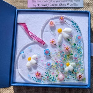 Puede incluir: Un adorno de vidrio en forma de corazón con diseños florales coloridos, presentado en una caja de regalo azul. El adorno presenta flores blancas, amarillas, rosas y azules con tallos verdes. Se adjunta una cinta rosa. El texto en la caja dice: "This handmade gift for you was lovingly made by Loxley Chapel Glass on Etsy."