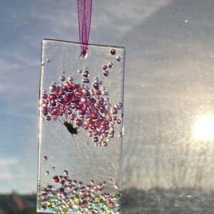 K&ouml;nnte beinhalten: Ein transparenter Glasanh&auml;nger mit einem rosa und violetten Blumendesign und schimmerndem Glitzer. Der Anh&auml;nger h&auml;ngt an einem violetten Band.