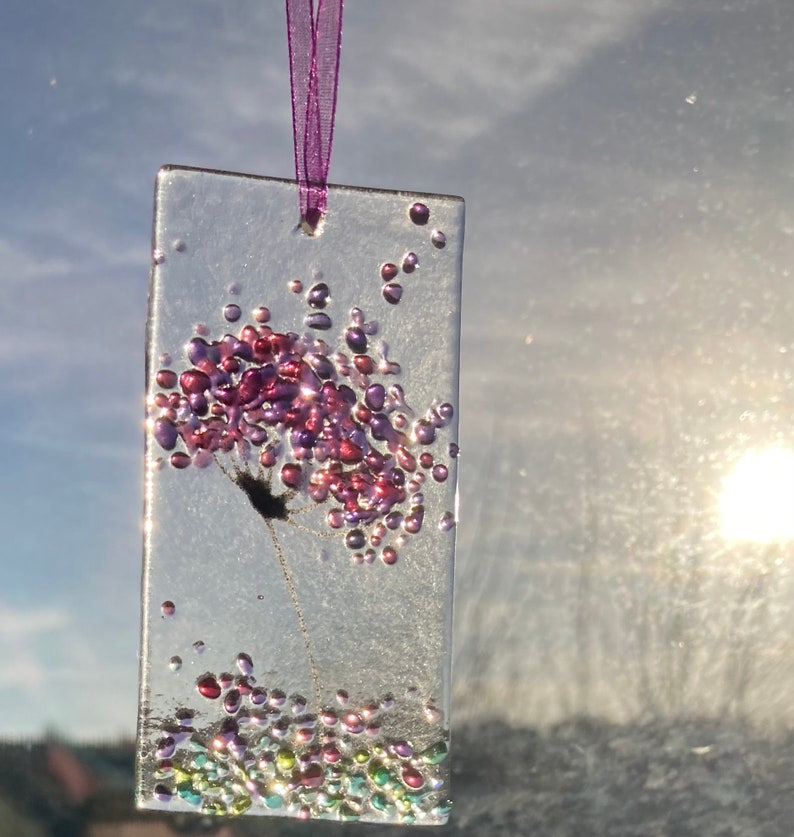 K&ouml;nnte beinhalten: Ein transparenter Glasanh&auml;nger mit einem rosa und violetten Blumendesign und schimmerndem Glitzer. Der Anh&auml;nger h&auml;ngt an einem violetten Band.
