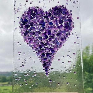 May include: A rectangular clear glass panel featuring a heart shape composed of numerous small, fused purple and violet glass pieces. The heart is surrounded by scattered purple glass fragments, and the panel is supported by a dark wooden base.