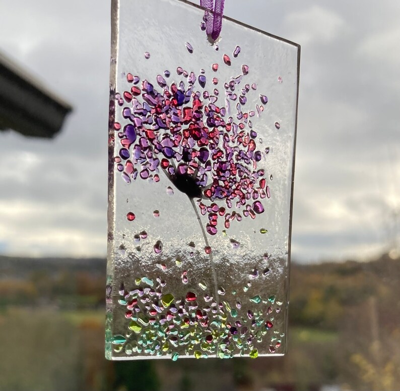K&ouml;nnte beinhalten: Ein Glasornament mit einer lila und pinkfarbenen Blume mit einem gr&uuml;nen Stiel. Die Blume besteht aus kleinen Glassplittern und ist von kleineren gr&uuml;nen, lila und pinkfarbenen Glassplittern umgeben.