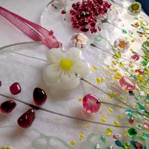 Op de afbeelding: Een handgemaakt glaskunstwerk met een witte bloem met een gele kern, omringd door kleurrijke glazen kralen en accenten. Een roze lint is bevestigd om op te hangen. Het ontwerp bevat rode, roze, groene en gele elementen.