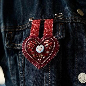 Puede incluir: Un corazón de fieltro rojo con un diseño de cuentas y una pequeña cara blanca en el centro. El corazón está sujeto a una chaqueta vaquera con un imperdible.