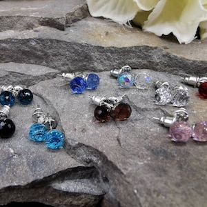 Op de afbeelding: Assortiment van kristallen oorbellen in verschillende kleuren, waaronder blauw, rood, roze, zwart en helder. De oorbellen worden gepresenteerd op een grijs rotsoppervlak.