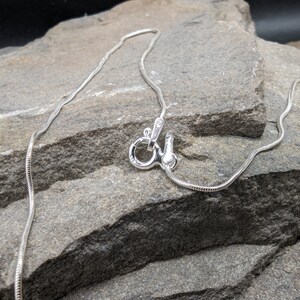 May include: A silver snake chain necklace with a lobster clasp. The chain is thin and flexible, with a smooth, reflective surface. The clasp is also silver and appears to be made of metal. The necklace is displayed on a grey stone surface.