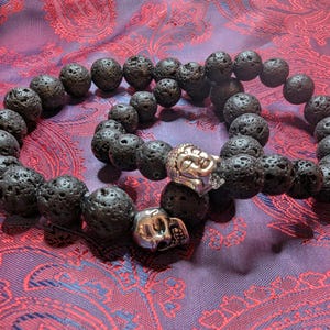 Puede incluir: Dos pulseras de cuentas de roca volcánica negra con dijes de calavera y cabeza de Buda de plata. Las pulseras están sobre una tela estampada de color morado y rojo.