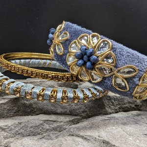Könnte beinhalten: Drei graue Stoffarmbänder mit goldenen Verzierungen und blauen und klaren Strasssteinen. Die Armbänder sind übereinander gestapelt.