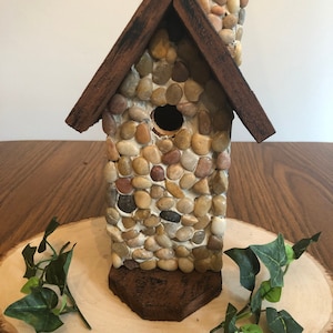 May include: A small birdhouse made of wood and decorated with stones. The birdhouse has a brown wooden roof and a chimney made of stones. The birdhouse is sitting on a wooden slice.