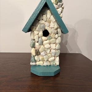 May include: A decorative birdhouse with a stone exterior and a teal roof and base. The birdhouse is made with small, smooth stones in shades of grey, beige, and green. The roof and base are painted teal.