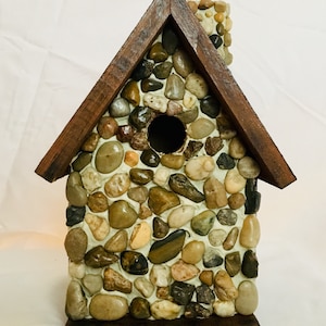 Könnte beinhalten: Ein dekoratives Vogelhaus mit Steinaußen und einem Holzdach. Das Vogelhaus ist aus unterschiedlich großen Steinen in Braun-, Beige- und Grautönen gebaut. Das Dach und die Basis bestehen aus dunkelbraunem Holz. Eine kreisförmige Öffnung ist sichtbar.