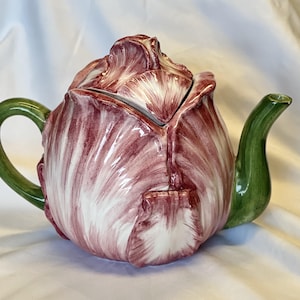 May include: A ceramic teapot shaped like a red and white flower with a green handle and spout. The teapot has a lid that fits snugly on top. The design is detailed and the colors are vibrant, making it a decorative kitchen item.