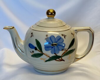 May include: Cream ceramic teapot with a blue floral design and gold detailing. The teapot has a rounded body, a curved spout, and a geometric handle. The lid is topped with a gold knob. The teapot is decorated with a blue flower.