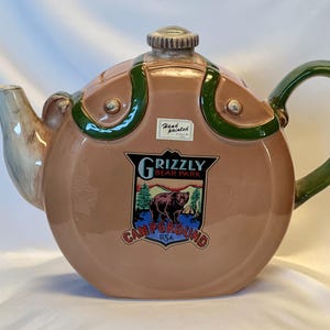 May include: A hand-painted ceramic teapot with a brown body, green handle, and accents. The teapot features a Grizzly Bear Park Campground USA emblem. The spout is light blue and the lid has a small knob.