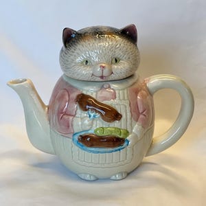 May include: A whimsical ceramic teapot shaped like a cat. The cat's head forms the lid, with a gray and white fur pattern. The body is pink and white, with a design of food on a plate. The teapot has a curved handle and spout.