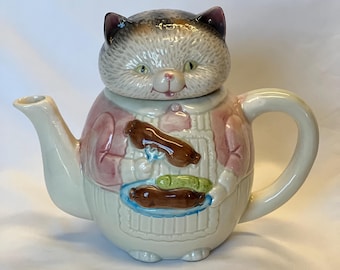 May include: A whimsical ceramic teapot shaped like a cat. The cat's head forms the lid, with a gray and white fur pattern. The body is pink and white, with a design of food on a plate. The teapot has a curved handle and spout.