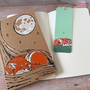 May include: A brown notebook and a green bookmark with a sleeping fox illustration. The notebook features a sleeping fox under a moon and stars design. The bookmark has a fox illustration on a green background.