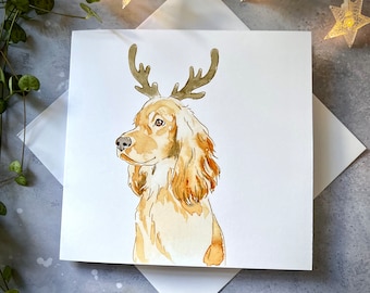 May include: A watercolor illustration of a golden cocker spaniel with reindeer antlers. The dog is looking to the right and has a light brown nose and brown eyes. The background is white.