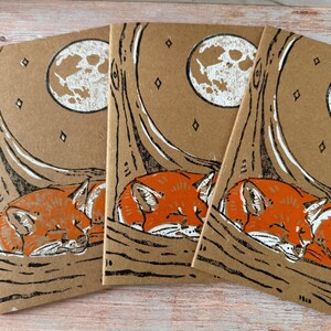 May include: Three notebooks with a nature-themed design. Each notebook features a sleeping orange fox under a tree with a full moon in the background. The design is printed in white and black on a brown paper background.