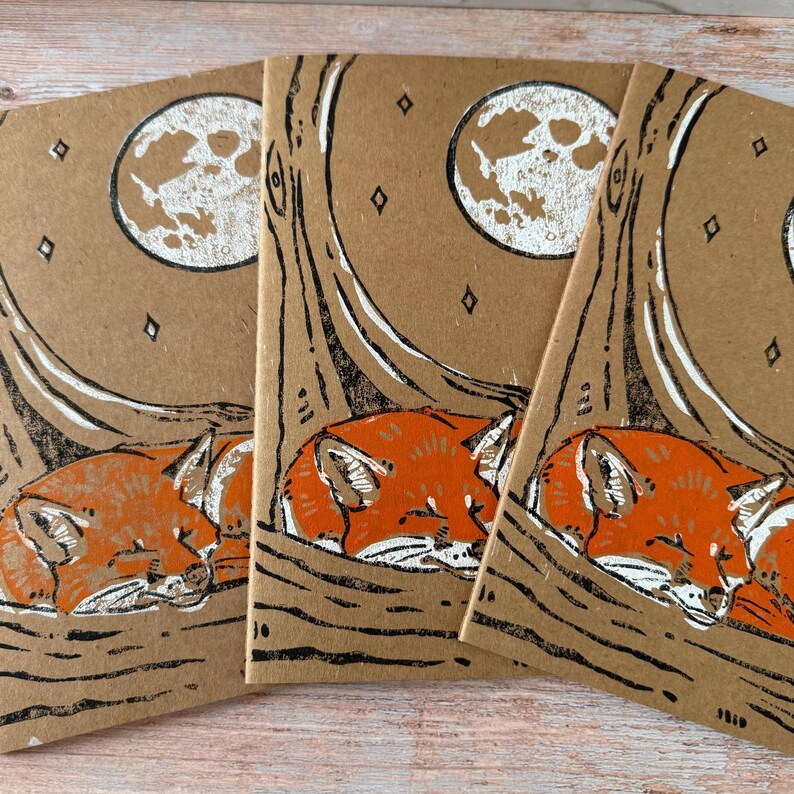 May include: Three notebooks with a nature-themed design. Each notebook features a sleeping orange fox under a tree with a full moon in the background. The design is printed in white and black on a brown paper background.