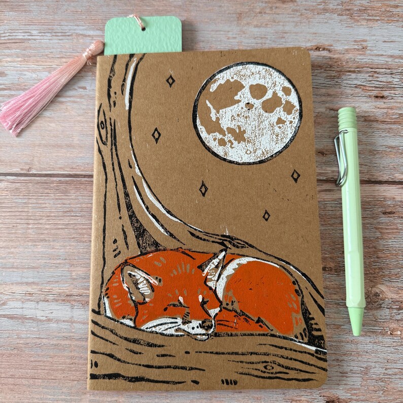 May include: A brown notebook with a sleeping fox illustration. The fox is orange and white, nestled under a tree with a full moon in the background. A light green pen and a pink tassel bookmark are included.