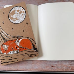 May include: A notebook with a brown cover featuring a sleeping fox illustration under a full moon. The notebook is open to reveal blank white pages. The fox is orange and white, and the moon is white against a brown background.