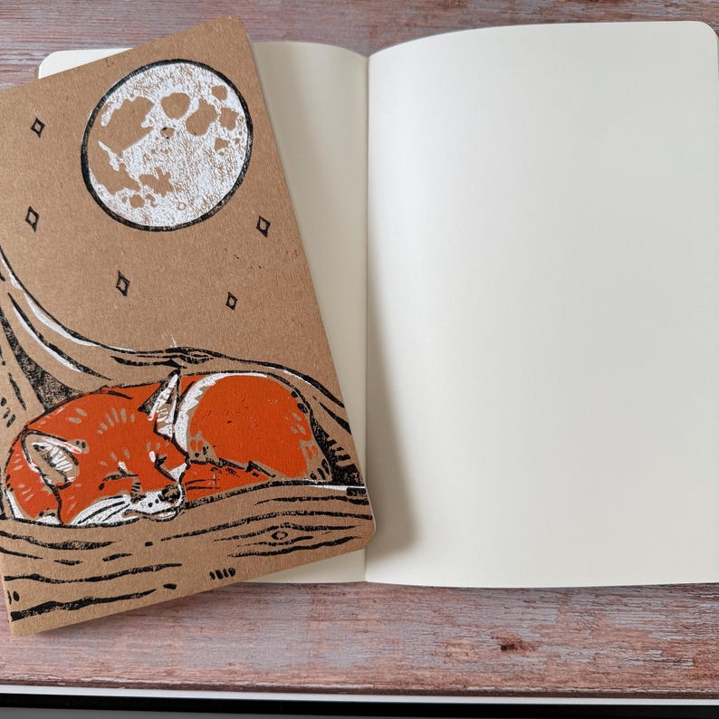 May include: A notebook with a brown cover featuring a sleeping fox illustration under a full moon. The notebook is open to reveal blank white pages. The fox is orange and white, and the moon is white against a brown background.