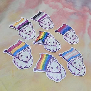 May include: A collection of glittery stickers featuring white bunnies holding flags with various LGBTQ+ pride flag designs. The flags include the asexual, rainbow, transgender, and other pride flags. The stickers have a white outline and are on a pink and white background.