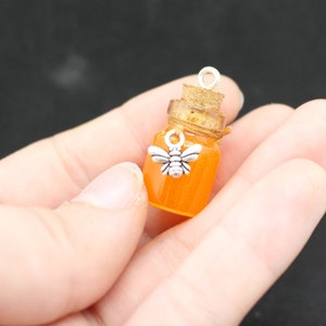 May include: A small glass jar filled with orange resin, resembling honey, with a cork stopper and a silver bee charm hanging from the top.