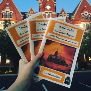 Puede incluir: Tres zines naranja y amarillo con el título "Spooky Stories about Insane Asylums...and how Mental Healthcare has Improved!"  Las zines se sostienen frente a un edificio de ladrillo rojo con una torre de reloj. Las zines son de @ReflectiveZines.
