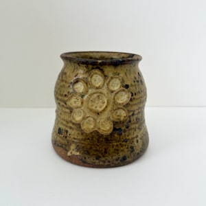 May include: A handcrafted ceramic vase with a textured, earthy appearance. The vase is primarily brown with darker speckles and a raised floral design. The opening is slightly flared, and the overall shape is rounded.
