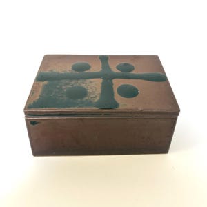 May include: A rectangular copper-toned metal box with a hinged lid. The top surface features a dark green enamel cross design with four circular accents, showing signs of wear and patina across the metallic finish of the container.