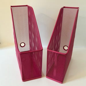 May include: Two magenta metal mesh magazine holders. Each has a rectangular base and a slanted back, with a circular hole near the bottom. The mesh design allows easy viewing of contents. Suitable for organising magazines, files, or documents.