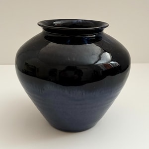 May include: A dark blue ceramic vase with a glossy finish. The vase has a rounded body and a slightly flared rim. The color transitions from a deep navy at the top to a lighter blue near the base. The vase is a decorative item.