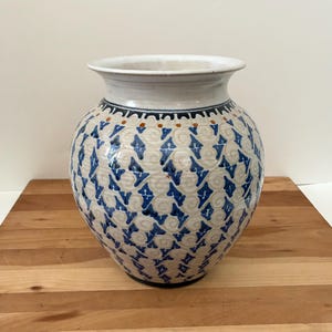 May include: A ceramic vase with a wide, rounded body and a flared rim. The vase features a cream-colored base with a repeating pattern of blue diamond shapes. The rim is white, and a dark blue and brown band encircles the neck.