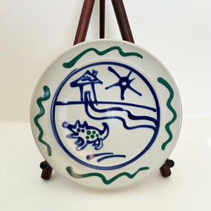 May include: A white ceramic plate with a hand-painted design featuring a house, sun, and dog in blue. The plate has a blue and green wavy border and is displayed on a wooden easel.