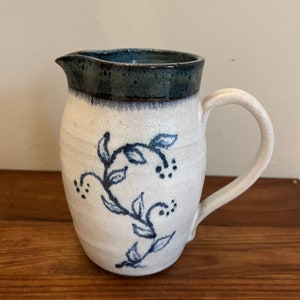 May include: A white ceramic jug with a blue floral design and a dark blue rim. The jug has a rounded body and a handle.