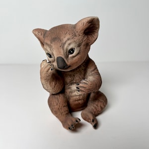 May include: A small, brown and black ceramic koala figurine sitting with its paws in front of it. The koala has a cute, innocent expression on its face.