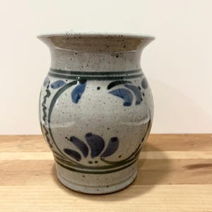 May include: A white ceramic vase with a blue and green floral design. The vase has a speckled glaze and a rounded body.
