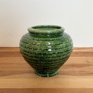 May include: A green ceramic vase with a round body and a small, flared base. The vase has horizontal, textured stripes in varying shades of green and black. The top rim is smooth and rounded. The vase is sitting on a wooden surface.