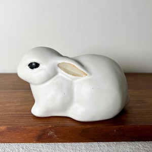 Może przedstawiać: Biała ceramiczna figurka królika z czarnymi oczami i jasnobrązowym uchem. Królik siedzi na drewnianej powierzchni.