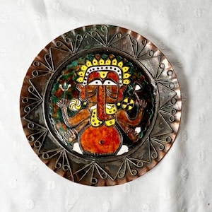 Peut inclure: Une assiette décorative en métal avec une illustration colorée d'une divinité à plusieurs bras et une tête d'éléphant. L'assiette a un bord de couleur cuivre avec un motif géométrique et une image centrale aux teintes rouges, jaunes et vertes.
