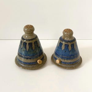 May include: A pair of bell-shaped ceramic salt and pepper shakers featuring a speckled tan base glaze with a blue drip pattern around the midsection. Each shaker has a small, round wooden plug inserted into the side near the base.