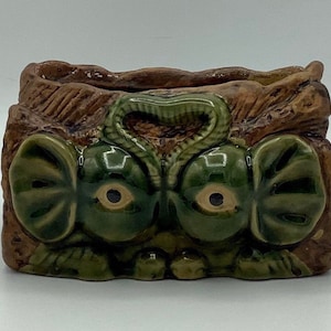 Könnte beinhalten: Ein Keramik-Pflanztopf in Form eines Gesichts mit grünen Augen, Ohren und einer braunen, strukturierten Oberseite. Der Topf ist so gestaltet, dass er wie eine Kreatur mit einem schlangenartigen Design auf der Oberseite aussieht.