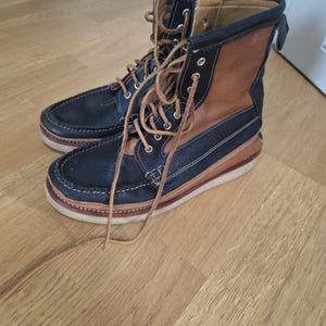 Puede incluir: Un par de botas altas que combinan paneles de cuero negro y marrón. Las botas tienen una puntera estilo mocasín, ojales metálicos, cordones de color tostado y una suela de goma gruesa de color claro con un detalle de costura visible en la base.
