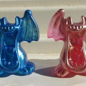 May include: Three small, iridescent dragon figurines in blue, pink, and purple. Each dragon has large wings and a smiling face. The figurines are sitting on a light-colored surface.