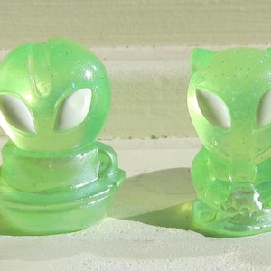May include: Five translucent green alien figurines with white eyes. The figures vary in design, with some standing upright and others in a seated or crawling position. The figures are displayed on a white surface.