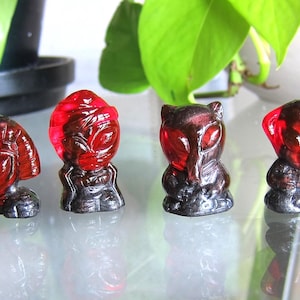 May include: A row of six small, alien-like figurines. Each figurine has a red, translucent head and a dark gray base. The figures vary in design, with some appearing to have wings or different head shapes. The figurines are displayed on a reflective surface.
